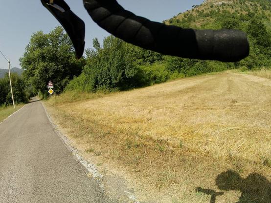 Strada di campagna che si infila tra gli alberi. In basso, una parte di campo con l'erba tagliata di recente e probabilmente convertita in una balla di fieno. Visibili alcuni elementi di una bicicletta, il lato destro del manubrio e la leva del freno.