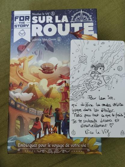 La boîte du jeu Sur la Route de Nicolas le Vif, illustré par Laëtitia Van Gasse. Posé sur la boîte, une dédicace sur une carte blanche avec un dessin au trait représentant une enfant avec des couettes et un cartable, montée à califourchon sur une fusée au milieu des astres.
Texte de la dédicace : Pour Lam Son, qui diffuse les ondes rôlistes jusque dans les étoiles. Merci pour ce que tu fais ! Je te souhaite drama et émerveillement ❤️
Nico Le Vif