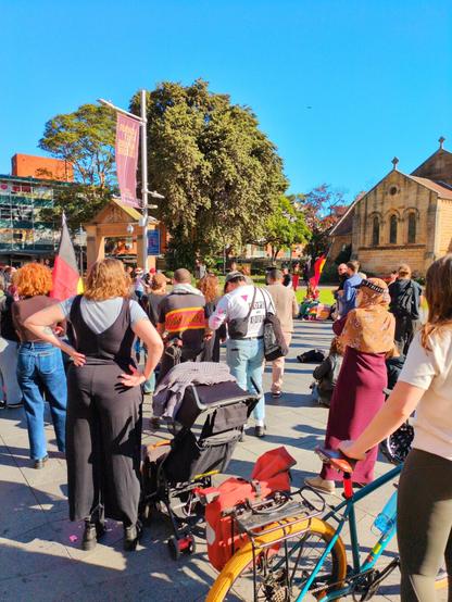 Aboriginal flags wave in front of a Cathedral. A multicultural crowd calls for justice.