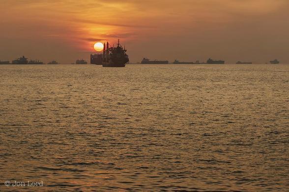 This Is A Colour Seascape Photo of a tropical sunrise and a long row of anchored merchant ship In Landscape Format. The Entire Image Is Tinted With A Golden Glow From A Just Risen Tropical Sun (Only A Few Degrees Above The Horizon). A Calm Sea Covers The Lower Three-Quarters Of The Frame With A Crisp Horizon Dividing The Ocean From The Sky. The Water Has Numerous Ripples Disturbing The Surface But Is Essentially Calm. Stretching Across The Horizon Is An Anchorage With A Great Many (Twenty-One In View) Cargo Ships At Anchor. The More Distant Vessels Are At Right Angles To The Camera Though The Two Closest, Above And To The Left Of Centre, Are A Medium Sized Container Ship And Car Carrier Are Nearly Parallel To The Lens. Just Above These Two Ships Is The Golden Rising Sun That Has Cast A Glow Across A Thin Band Of High Level Cirrus Clouds. 
The Location: The Singapore Straits, South China Sea (2017).