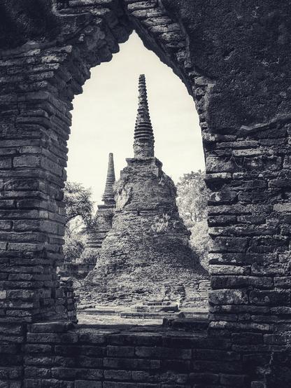 a view through ancient and narrow window of a partially collapsed palace wall of Ayutthaya, the ancient capital of Siam, frames 2 iconic stupa structures -- bell shaped shrine / meditation areas