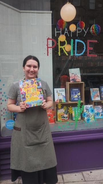 Bookseller Sarah holding a copy of A Year of Pride and Joy outside the Pride display window of the Edinburgh Bookshop