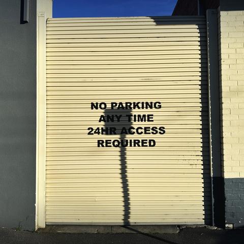 A cream-coloured corrugated garage door that says “no parking any time 24 hour access required” in the middle. Also in the middle
Is the shadow of a nearby street sign.