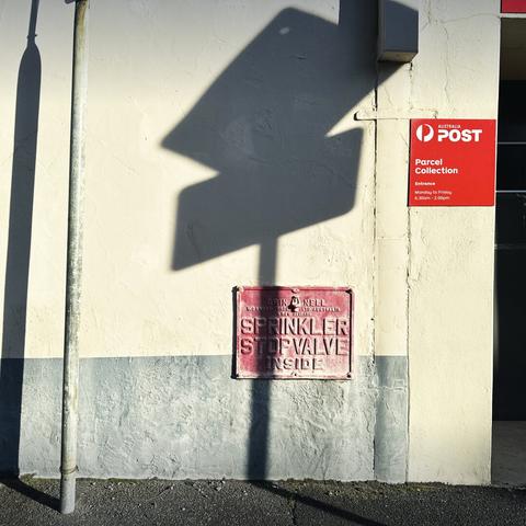 An old, red sprinkler stop valve sign attached to a wall, with a diamond + letter - shaped shadow of a nearby street sign falling across it in the morning sun. A red Australia Post sign is over on the right at the edge of the frame.