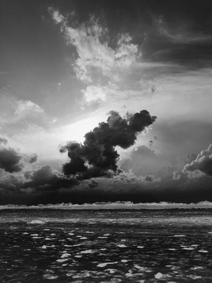 Dramatic black and white photo of the setting sun behind some storm clouds over a sandy beach.