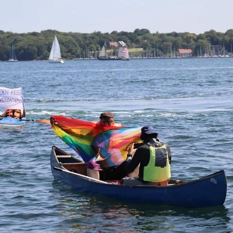 Foto: Ruderboot mit zwei Personen von hinten. Die vordere hat eine Regenbogenfahne auf dem Rücken und mit den Armen gespreizt.
