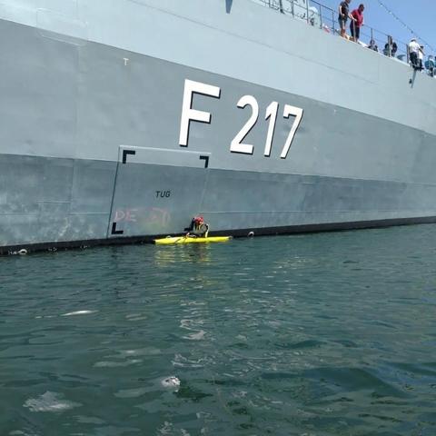 Foto: die Hälfte des Bildes wird von einem Kriegsschiff "F217" gefüllt, die andere ist Wasser. Ein kleines gelbes Kajak ist am Schiff zu sehen.