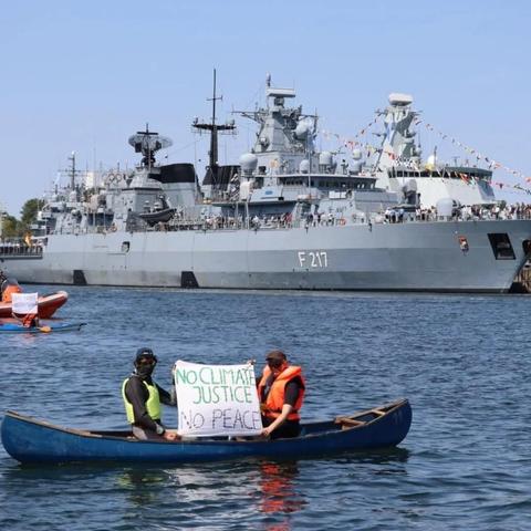 Foto: Im Hintergrund zwei große Kriegsschiffe (F217, ?). 
Im Vordergrund eine Ruderboot mit zwei Personen in Schwimmwesten (gelb + orange), die eine Transpi zwischen sich halten: "No climatejustice, no peace"