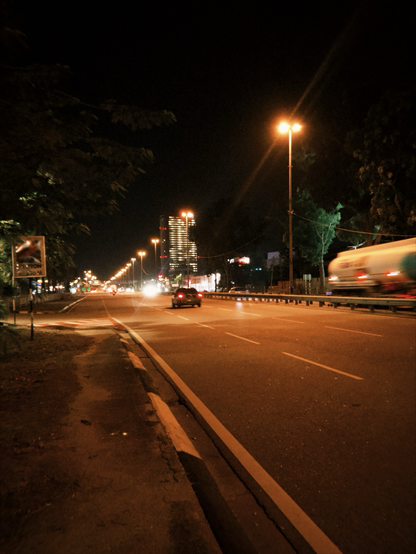 The image depicts a nighttime urban street scene. The road is well-lit by streetlights, with a series of bright yellow lights lining the sides. The road has multiple lanes, with visible lane markings. A car is seen in the center lane, moving towards the camera, with its headlights on. On the right side of the road, a large truck is captured in motion, creating a blur effect due to its speed. The background features a tall building with illuminated windows, suggesting an urban environment. The sky is dark, indicating it is nighttime. On the left side of the road, there is a sidewalk with some trees and a signpost, partially visible. The overall atmosphere is quiet, with minimal traffic, and the scene is illuminated by artificial lighting.
