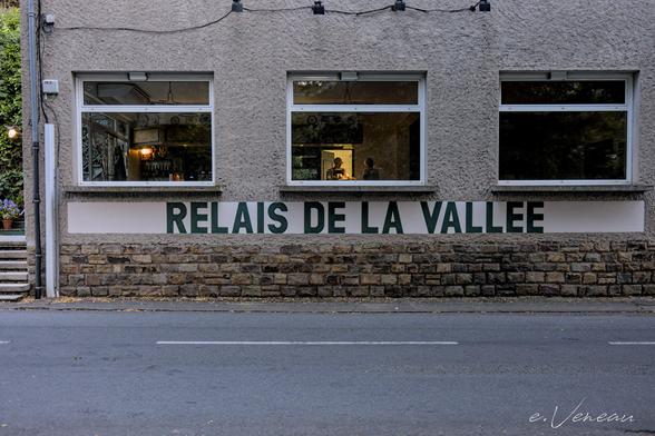 The front of a roadside restaurant. Through the windows, whose panes reflect the foliage, we can make out 2 men facing each other at the bar.