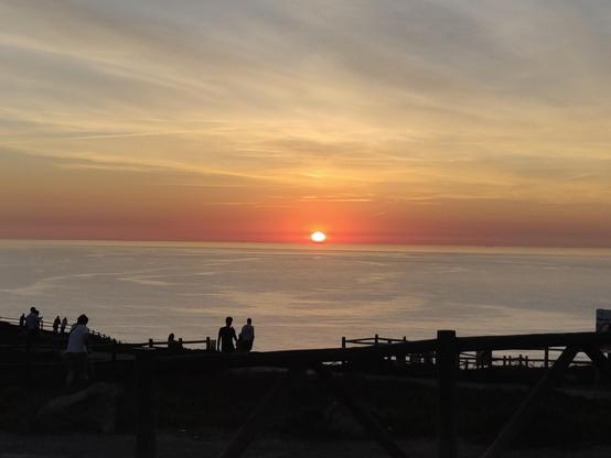 A imagem captura um pôr do sol sobre o mar, com o sol meio escondido no horizonte longínquo, uma semi-esfera, quase um ponto de luz amarelo-vivo com uma difusa auréola laranja  que se espalha em tonalidade mais escura ao longo da linha do horizonte, reflectindo uma luz amarelada difundida no céu nublado, cinzento e branco. O mar chão é de cor cinza azulado e parece reflectir o céu nublado.Em primeiro plano, e na escuridão, são  visíveis as silhuetas em contra-luz de uma cercadura rústica de madeira  e duas pessoas de pé, viradas para o sol poente no que parece ser a beira de uma falésia. A cena é serena e evoca uma sensação de paz e contemplação.