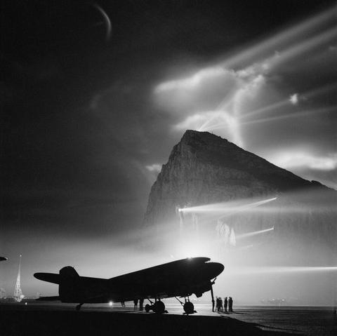 Black and white photo of a Douglas Dakota aircraft, silhouetted by searchlights on the Rock of Gibraltar during World War II.