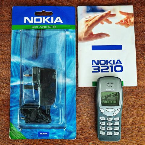 Photo of an old Nokia 3210 on wooden table, powered on showing the boot screen of hands touching. A manual is above it and an unopened official Nokia charger to the left