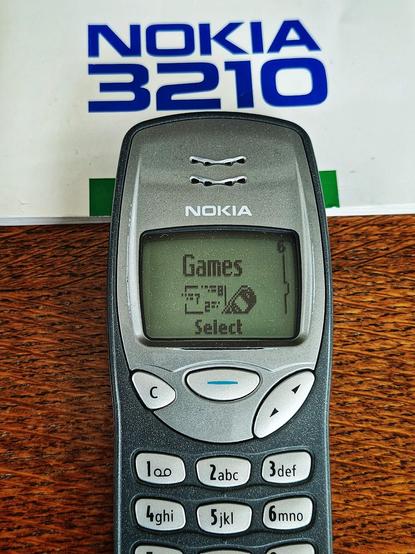 Close-up photo of an old Nokia 3210 on wooden table, powered on showing the "Games" menu screen. A manual is above it with the phone model title.