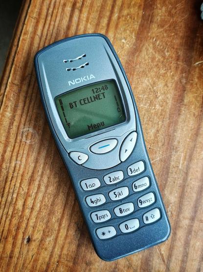Photo of an old Nokia 3210 on wooden table, powered on showing the home screen and "BT CELLNET" network logo. NOTE: This logo shows for about 1 second before renaming to "giffgaff" (my SIM/network provider).