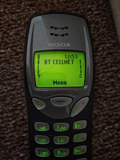 Close-up darker photo of an old Nokia 3210 a carpet, powered on with the backlight glowing while charging, showing the home screen and "BT CELLNET" network logo. NOTE: This logo shows for about 1 second before renaming to "giffgaff" (my SIM/network provider).