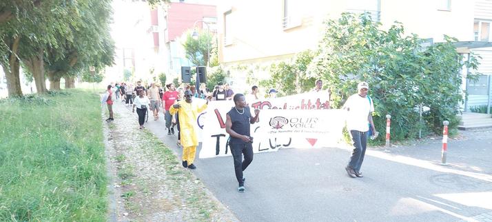 Demonstration in Freiburg zum Weltgeflüchtetentag. Viele Menschen laufen mit Transparenten und einem kleinen Lautsprechersystem eine Straße entlang. Die Sonne scheint hell. Auf dem vordersten Transpi ist "Our Voice" zu erkennen.