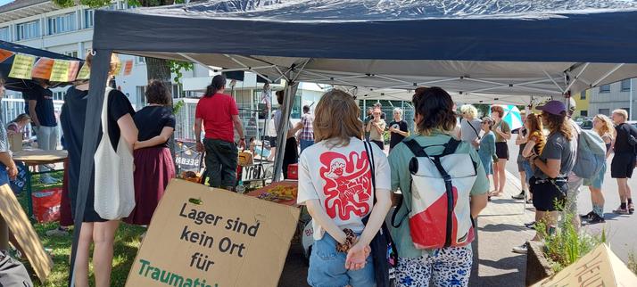 Demonstration vor der LEA (Landeserstaufnahme) in Freiburg. Es sind viele Menschen von hinten zu sehen, etliche stehen unter einem Pavillion. Vorne steht ein Schild, auf dem steht: "Lager sind kein Ort für Traumatisierte Menschen".