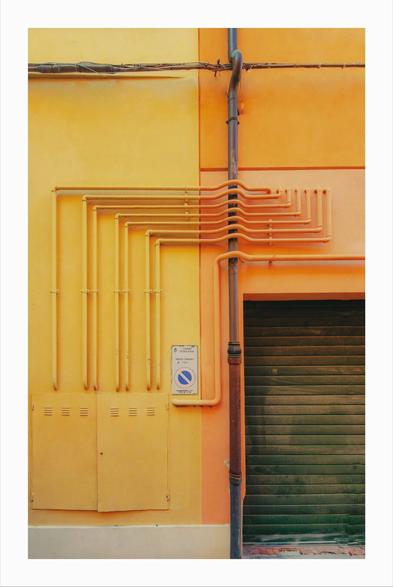This photo is a section of a building's exterior wall. The wall is divided into two color sections - there is a pale yellow on the left and a slightly more saturated orange on the right. Several parallel yellow pipes run horizontally across the left section, while a darker, rusty-colored vertical drainpipe runs down the center, connecting the two color sections. There's a green roll-up garage door to the right. A small Italian traffic sign is affixed to the wall in between the pipes and the door.