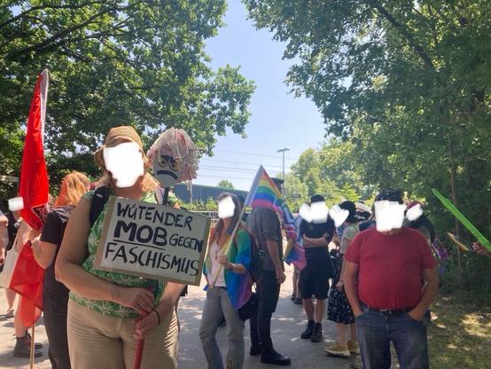 Demonstrierende mit roten Antifa- und bunten Pride-Flaggen; ein Mensch hält einen Wischmob mit aufgemaltem Gesicht und einem Schild mit dem Text „Wütender Mob gegen Faschismus“