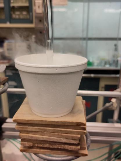 A Styrofoam cup sits on at least 6 wooden rectangles, hastily stacked on a ring stand clamped to an aluminum frame. The cup smokes with boiling liquid nitrogen, and the cold cloud falls behind the cup. A clear glass ampoule is suspended into the center of the cup with frost around the outside. A small clear tube is visible within, and a blurry fume hood with samples and reagents is visible behind the cup.