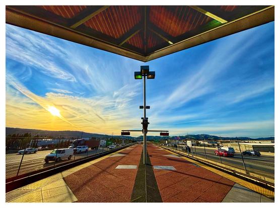 Landscape-orientation photo of the open-air station platform for the BART train in Dublin, California. The roof overhang and the platform surface both form visual triangles at the top and bottom of the image, as they recede towards a signal post standing at the center. Other symmetrical geometric patterns are formed by the beams and rows of parallel lines on the underside of the overhang, as well as the variously configured tiles on the platform, which are predominantly in yellow and reddish brown. Freeway traffic flows on either side of the platform. A line of low mountains sits at the horizon. The vivid blue sky is streaked with white cirrus clouds and washed in golden light at left frame by the afternoon sun.