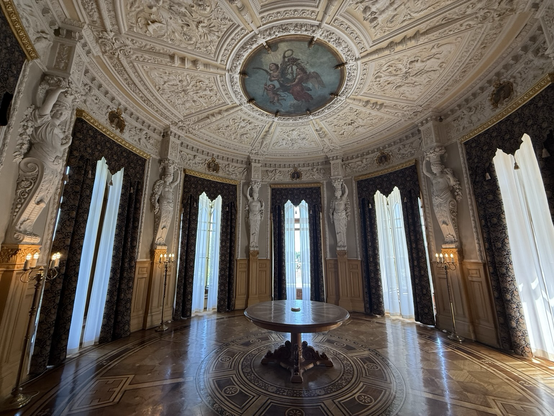 A circular room, highly decorated with ornate plasterwork to the ceiling and tall windows around the room. A single circular table sits in the middle.