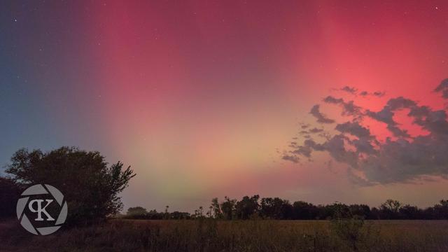 A photo of northern lights over a field in northeast Oklahoma. The lights are vibrant curtains of red and pink, with some shades of green visible closer to the horizon. The field is cast in an ethereal glow by the auroral light, with trees and a distant treeline visible in silhouette.