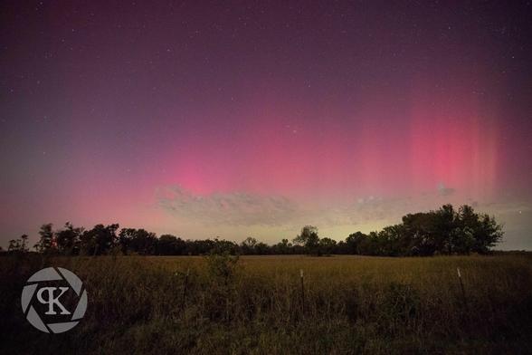 A photo of northern lights over a field in northeast Oklahoma. The lights are vibrant curtains of red and pink, with some shades of green visible closer to the horizon. The field is cast in an ethereal glow by the auroral light, with trees and a distant treeline visible in silhouette.
