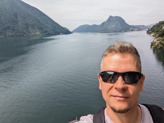 Author's face, a bit red from the sun, in wide sunglasses. In the back there's a wide body of water, filling a glacial valley, with almost tropical greenery on the sides. The air is somewhat hazy.