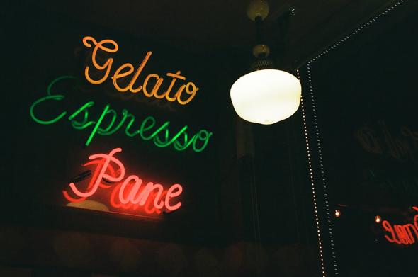 Neon yellow, green, and red sign that reads, “Gelato, Espresso, Pane” in Italian. There are diner lights that shines beside it.