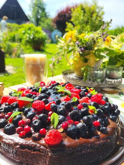 Das Bild zeigt eine dekorative Torte mit einer glänzenden Schokoladencreme, die mit einer Vielzahl von Früchten wie Blaubeeren, Raspeln und roten Beeren verziert ist. Die Torte ist auf einem Teller mit einer dekorativen Metallplatte platziert, die auch zwei Gläser enthält. Im Hintergrund ist ein sonniger Garten mit grünen Pflanzen und Blumen zu sehen, darunter ein Blumenstrauß in einem gelben Topf. Ein Glas mit einem Getränk steht auf dem Tisch. Die Szene wirkt idyllisch und einladend, ideal für einen sonnigen Tag im Freien.