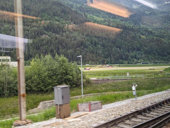 Ambri Airport in Ticino valley, seen from a passing train. As the airport is taking up a significant part of the valley and doesn't see that much traffic, there's a road cutting across it. A tractor transporting a bale of hay can be seen crossing the airport's runway.
