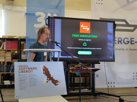 Filippo Della Bianca presenta la sua slide sul "Fedi Mercatino". Sullo sfondo c'è uno striscione del MERGE-it. In primo piano un cartellone su cos'è il software libero, fornito dalla sede.