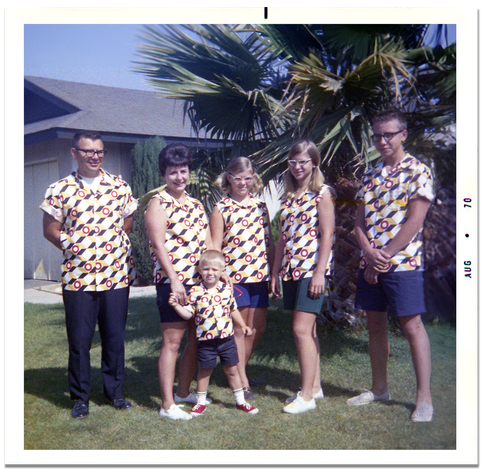 A family of six wears matching shirts with a loud, 3-d pattern. They stand for a portrait in the front yard of a suburban home in California. There is a garage and palm tree behind them.