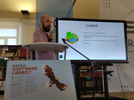 Zughy, serissimo, guarda il pubblico fulminandolo (positivamente) con lo sguardo, mentre mostra Luanti: software libero meraviglioso, con foglio della presentazione annesso, dettagliandone le caratteristiche di estetica, economicità e modularità.