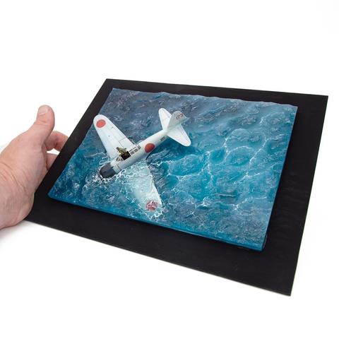 The image presents an extraordinary miniature diorama that masterfully captures a dramatic yet serene scene of a Japanese WWII-era plane sinking into a turbulent sea. The centerpiece of this diorama is a meticulously detailed model of what appears to be a Mitsubishi A6M Zero, a famous Japanese fighter aircraft. The plane’s iconic white body is accented by vivid red rising sun insignias on its wings and fuselage, characteristic of Japan’s wartime markings. The level of detail on the plane is remarkable, from its slightly weathered paint suggesting prolonged use to the tiny pilot figure seated in the open cockpit, frozen mid-action.