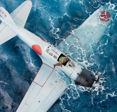 This close-up view intensifies the drama of the diorama, centering on the sinking Japanese WWII-era plane in a stormy sea. The white fuselage of the aircraft is brilliantly rendered, with subtle weathering and paneling that enhance its realism. The vivid red rising sun insignias, clean and bold, draw the eye immediately, grounding the model in its historical context. The cockpit, now partially open, reveals a pilot figure in a moment of critical action. Dressed in a faded green life vest and tan clothing, the pilot is captured mid-motion as he scrambles out of the sinking plane, his posture conveying urgency and resolve. The vivid colors of the ocean and the plane contrast strikingly, creating a visually arresting tableau of desperation and survival.