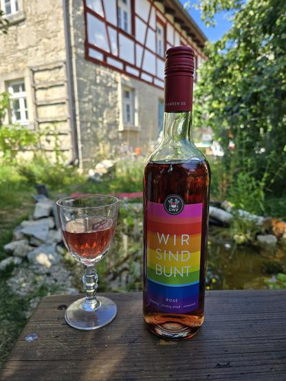 Das Bild zeigt eine Flasche rosé Wein mit einem bunten Etikett, das die Worte "WIR SIND BUNT" in verschiedenen Farben zeigt. Die Flasche steht auf einem Holztisch vor einem traditionellen Fachwerkhaus mit Steinmauern. Neben der Flasche steht ein Weinglas, das halb gefüllt mit rosé Wein ist. Im Hintergrund ist ein Garten mit Grünzeug und ein kleiner Teich zu sehen. Die Flasche trägt den Aufdruck "Franken EG" und "GWF" auf dem Etikett. Die Farben des Etiketts sind in den Regenbogenfarben angeordnet, was auf eine Botschaft der Vielfalt hinweist.