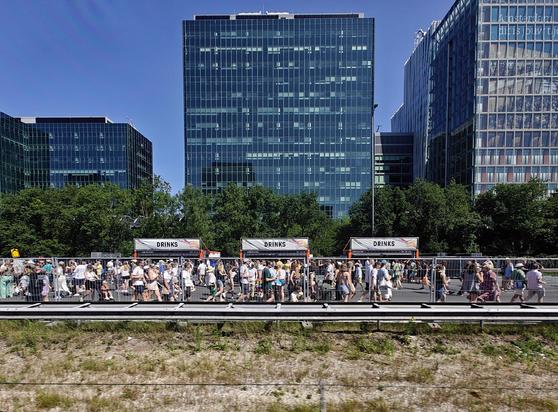 A large crowd of people walking on a closed motorway, passing in front of three beverage stands labeled "DRINKS". In the background one can see modern glass office buildings.