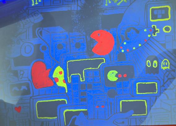 A postcard with a stamped UV-fluorescent pattern with visible highlighting of some elements. The UV pattern is wild mix of game figures, computers, a "WASD" key set, a cat in a UFO, cute ghosts. Highlighted in neon colors are some ghosts, pacman, computer screens and electronic connections.