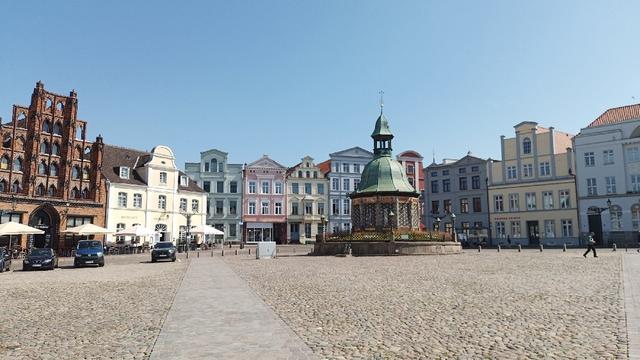 Blick auf die südöstliche Ecke des rd. 1ha großen Wismarer Marktplatzes. Kopfsteinpflaster, farbige Giebelhäuser aus unterschiedlichen Jhd. vom Mittelalter bis heute, darunter ein rotziegelgotisches Kaufmannshaus (Alter Schwede), das Reuterhaus u.a. In der Marktecke die Wasserkunst, ein Zisternen und Pumpenhaus aus dem 16. Jhd.. Blauer Himmel über und wenige Leute auf der nachmittags schattenlosen Seite des Platzes.
