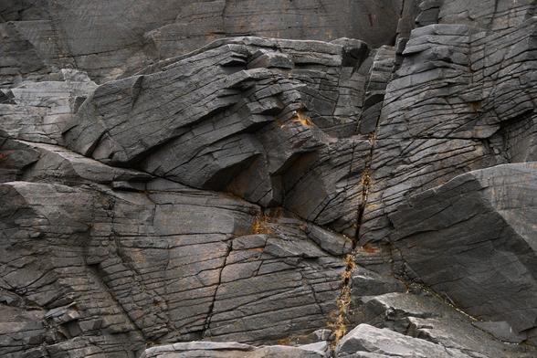 An abstract-ish photo of large sea stack rocks on a beach. The rocks are dark grey with fracture lines running mostly parallel and changing depending on the shape of the rock.