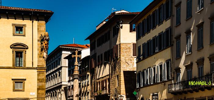 Sunlit Florence streetscape showing yellow-tinged stucco buildings with dark green shutters, a tall marble column crowned by a cross in the centre, and clear blue sky overhead.
