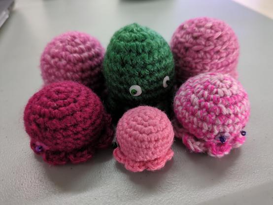 Photo de six petits poulpes au crochet posé sur une table, cinq d'entre eux sont roses, dans des nuances différentes, le dernier est vert