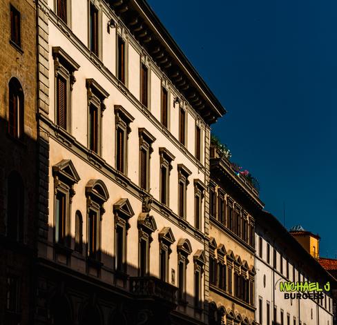 Tall Renaissance-style building in Florence lit by low sun, with cream walls, grey stone trim, wooden shutters, patterned shadows, and a rooftop garden against a deep blue sky.