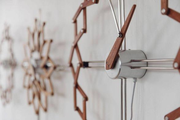 Close-up shot of a unique wall clock featuring a geometric design with wooden frames, set against a light textured background. Two other versions of the same clock are blurred out in the background.