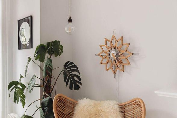 A cozy corner features a rattan chair with a fluffy cushion, a wooden star-shaped clock, a hanging light bulb, and lush green plants.