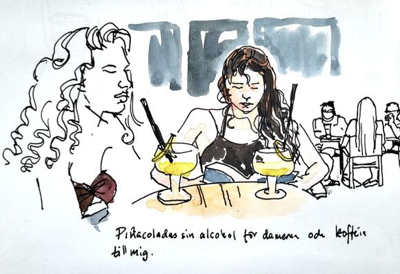 Två unga damer dricker pinacolada. Två glas på fot med långa svarta sugrör och citronskivor. Den ena läser en bok. I bakgrunden sitter en grupp människor vid ett bord.
