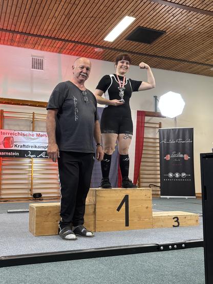 Woman standing on a podium marked 1, holding a trophy with her right hand and flexing her bicep with her left arm. A man stands to her right.